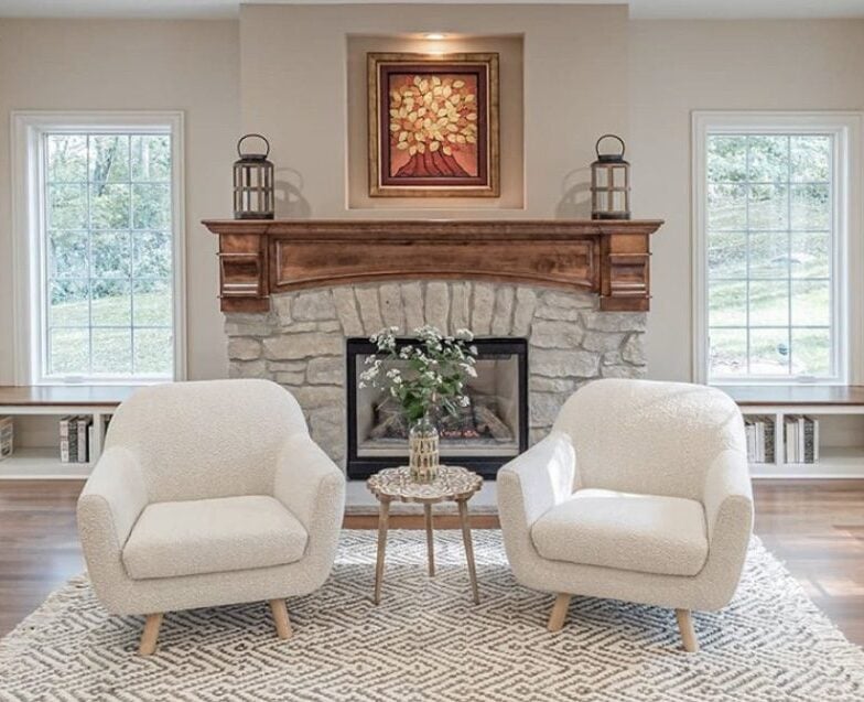 Stone fireplace with a wooden mantel behind two chairs and a table with a flower.